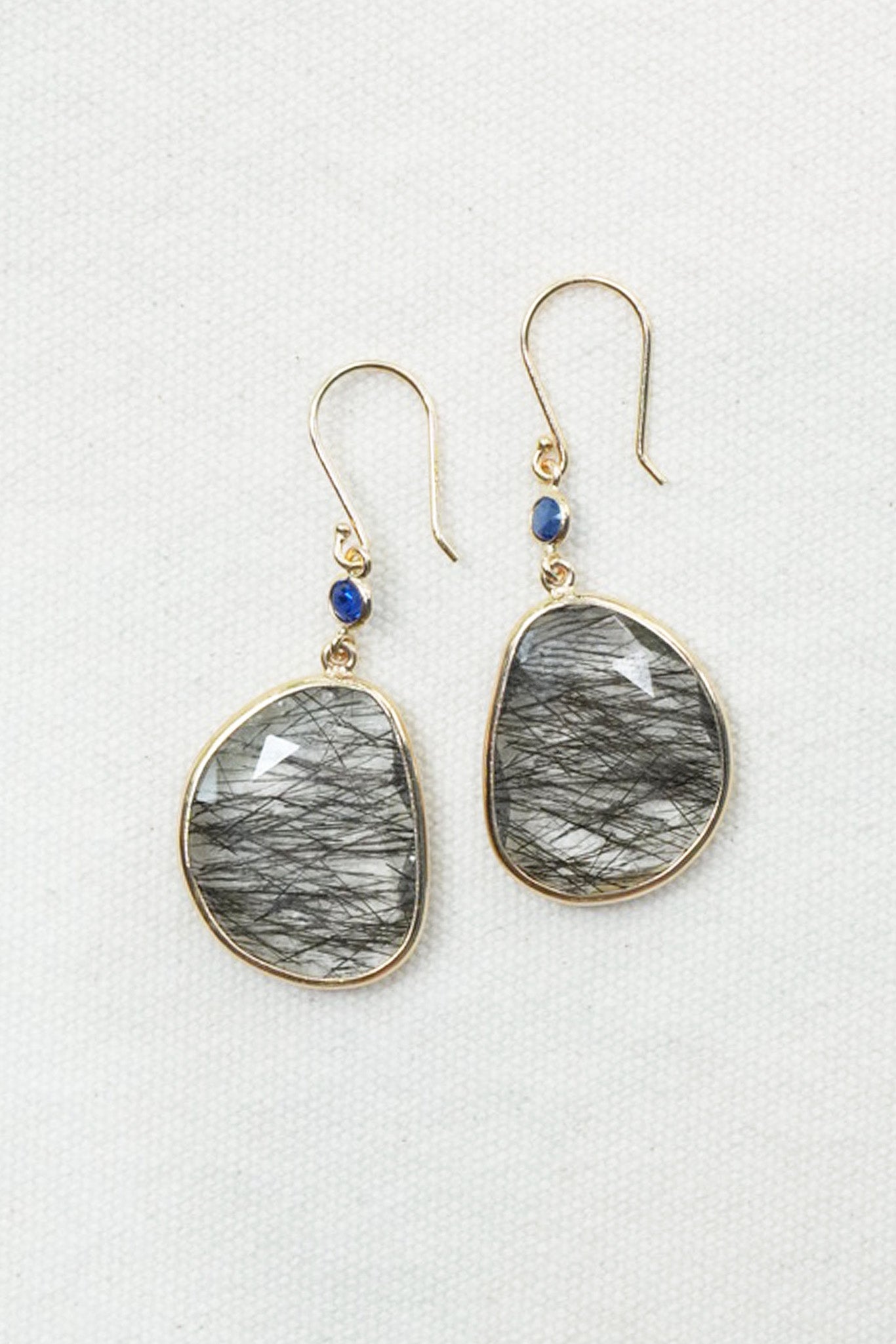 A pair of gold-tone dangle earrings with irregular teardrop-shaped gray and black hair-on-hide stones, accented by small round blue beads, shown on a textured white background.