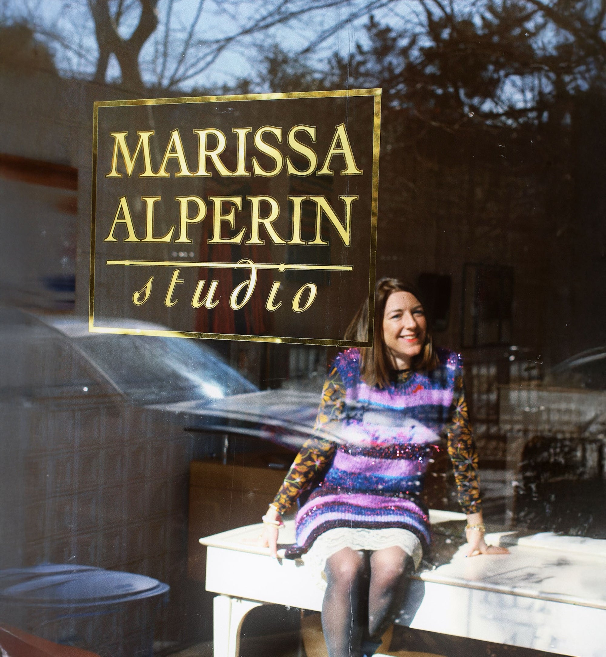Marissa sitting on a bench in front of her studio glass door with 'Marissa Alperin Studio' sign.