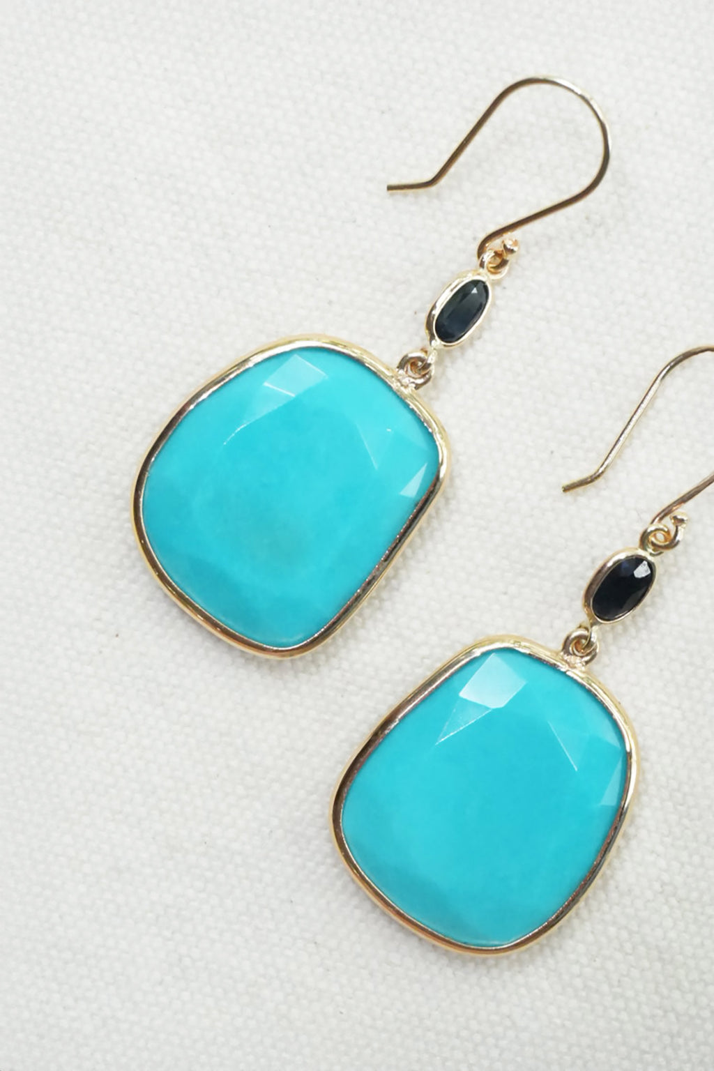 A pair of gold-tone drop earrings featuring large, rounded turquoise-blue cabochon stones and small black oval accents, displayed on a textured off-white fabric background.