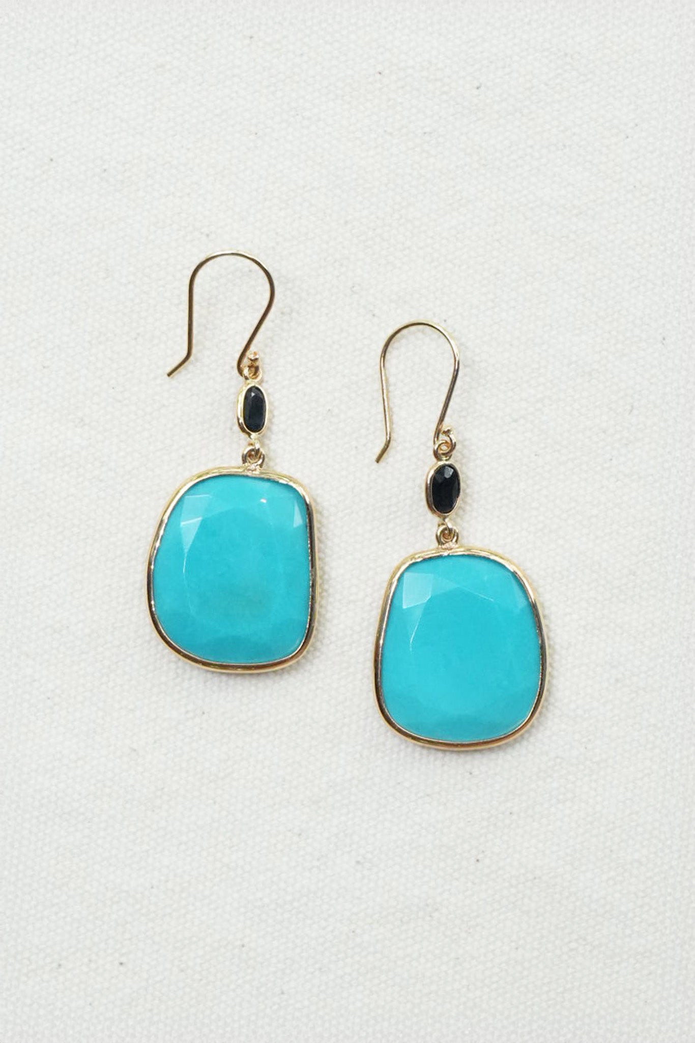 A pair of gold-tone drop earrings featuring large, rounded turquoise-blue cabochon stones, accented with small black oval beads, displayed on a textured off-white background.