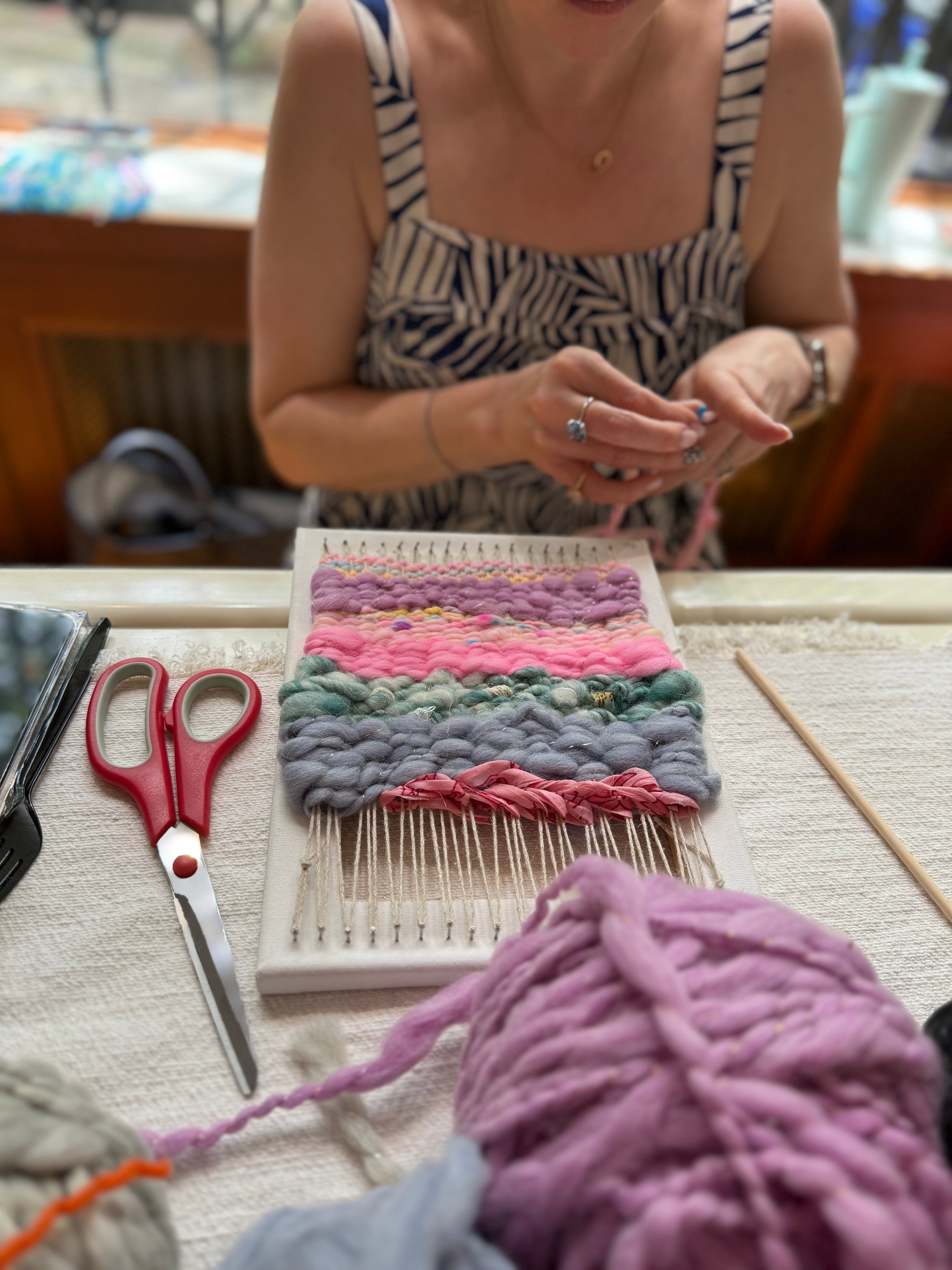 Person working with yarn and a loom on a table with scissors and other materials.