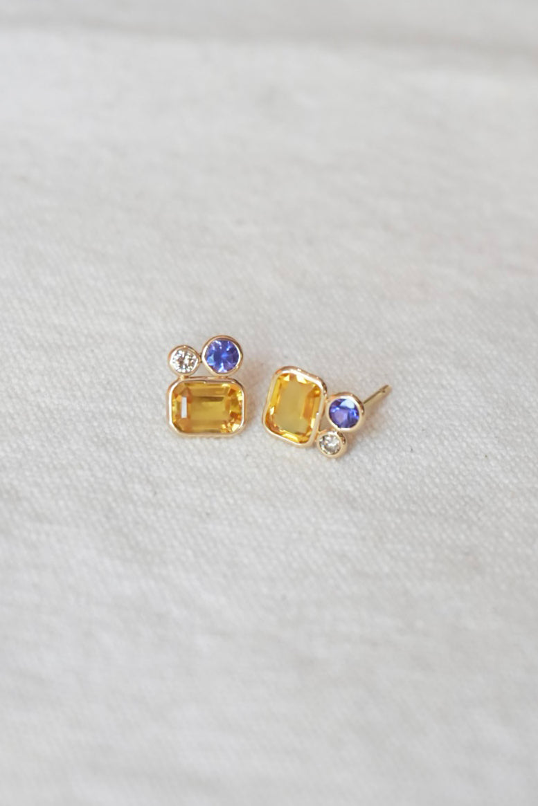 Gold stud earrings with yellow and purple gemstones on a white fabric background