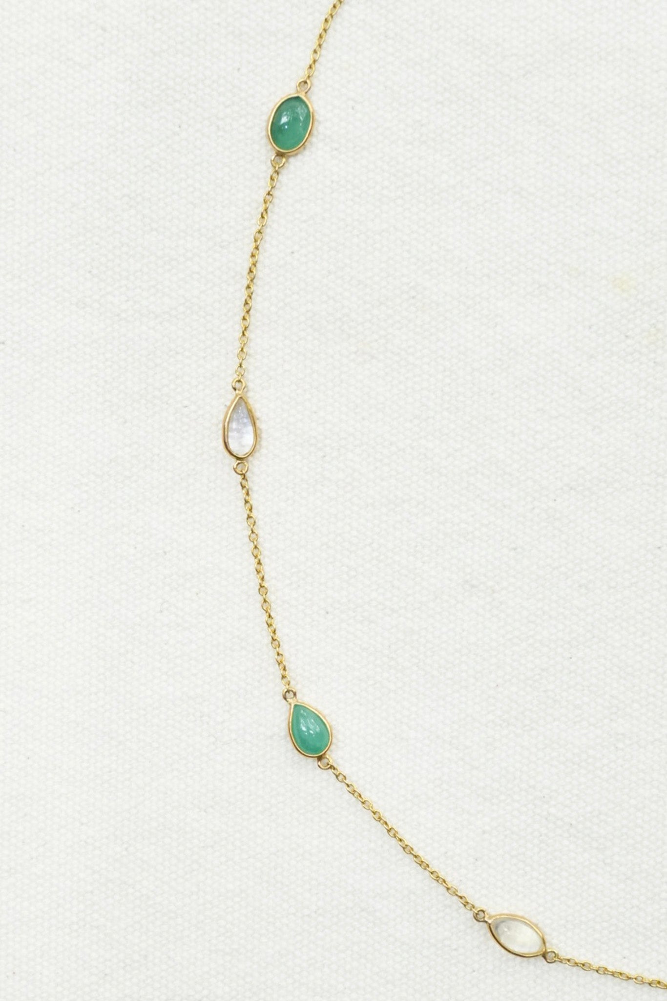 A delicate gold chain necklace featuring three green teardrop-shaped stones and two white teardrop-shaped stones, laid out on a textured off-white background.