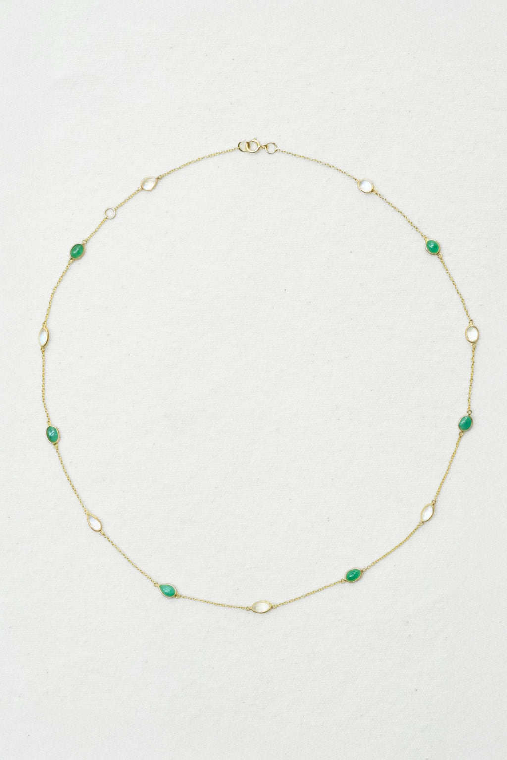 A delicate gold chain necklace featuring small, evenly spaced green oval gemstones, laid out in a circle on a textured off-white background.