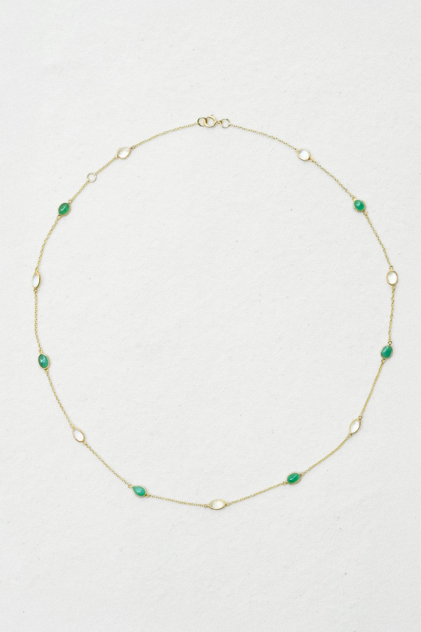 A delicate gold chain necklace featuring small, evenly spaced green oval gemstones, laid out in a circle on a textured off-white background.