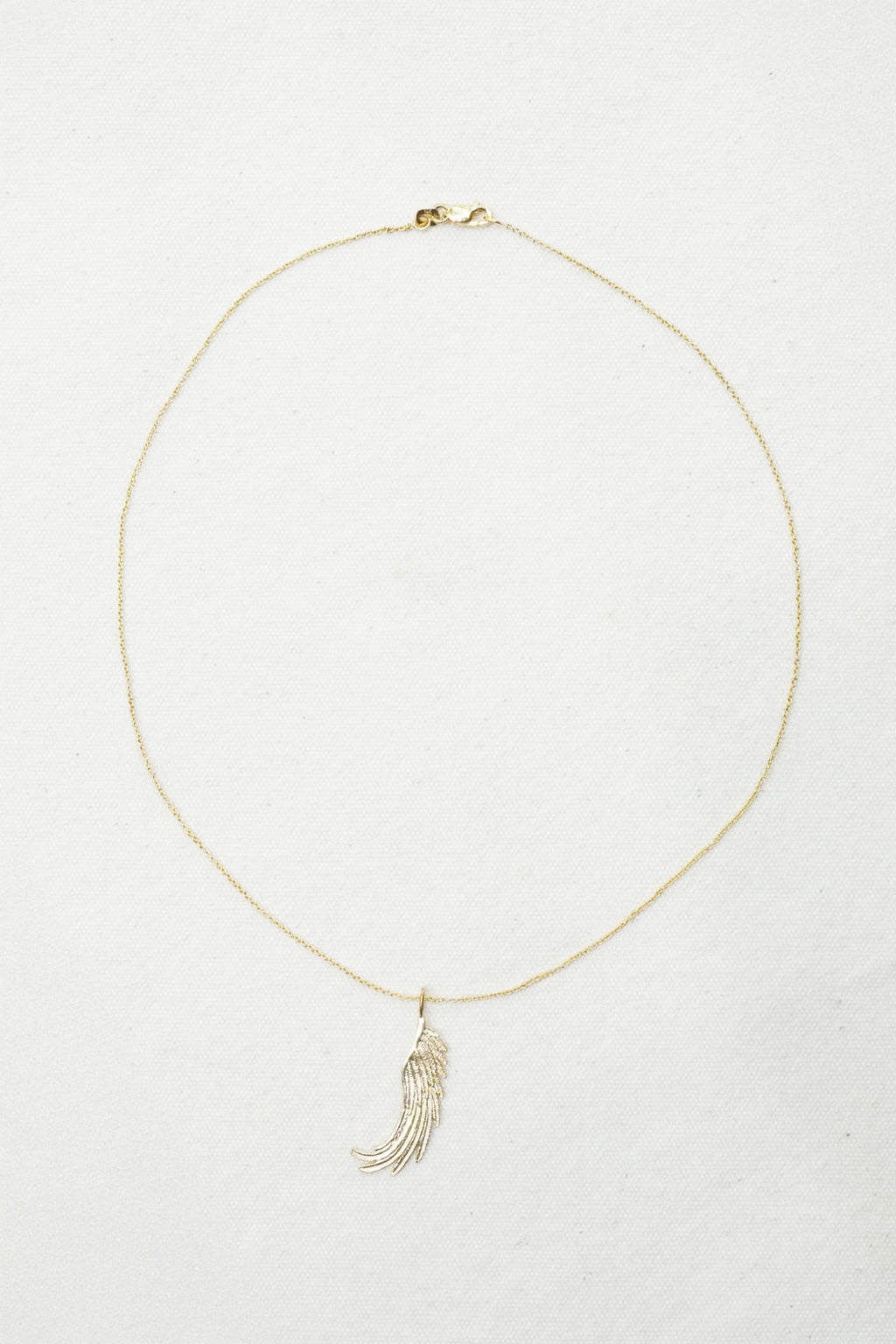 A delicate gold chain necklace with a small feather or wing-shaped pendant, displayed on a textured off-white background.
