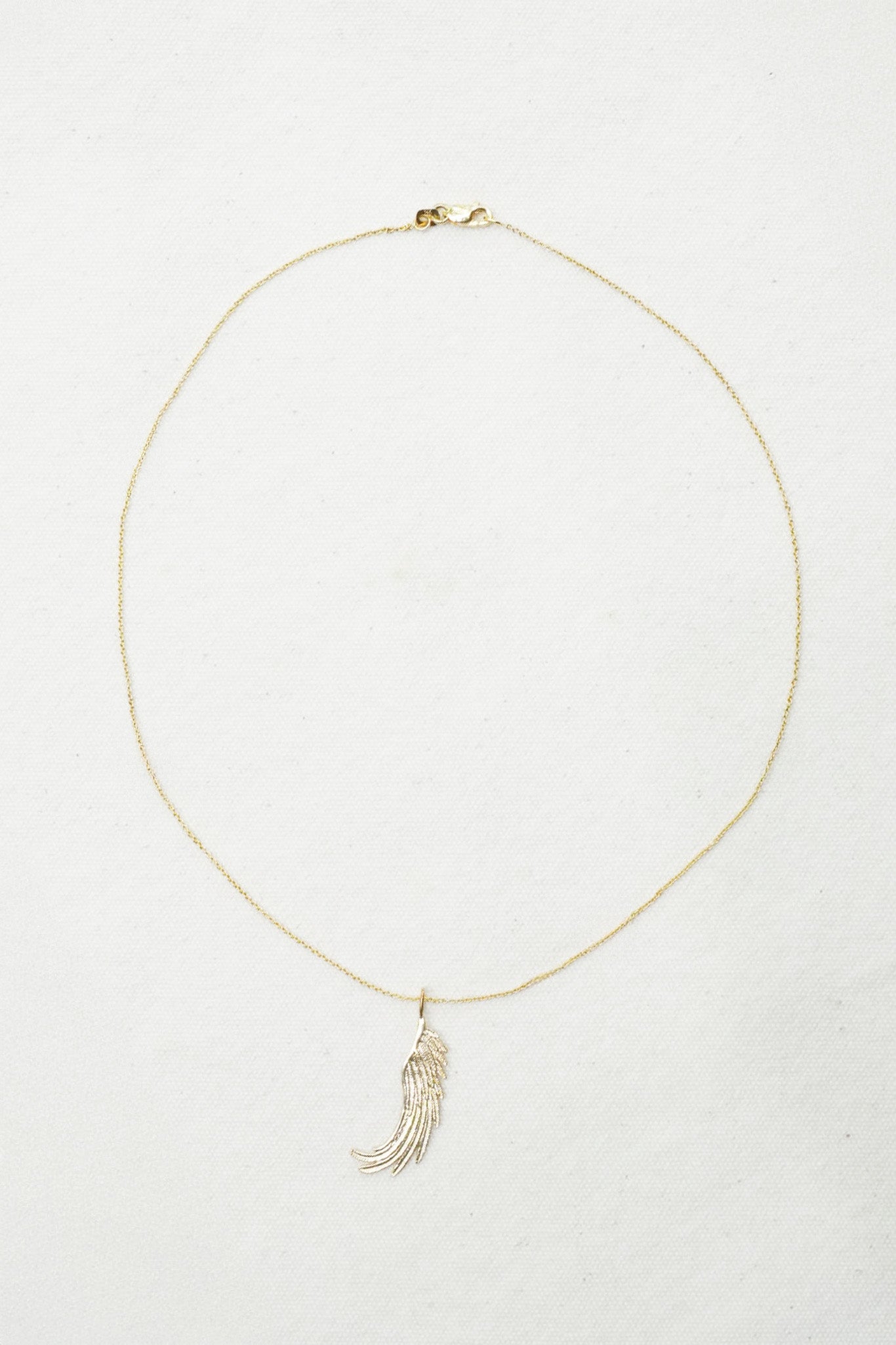 A delicate gold chain necklace with a small feather or wing-shaped pendant, displayed on a textured off-white background.