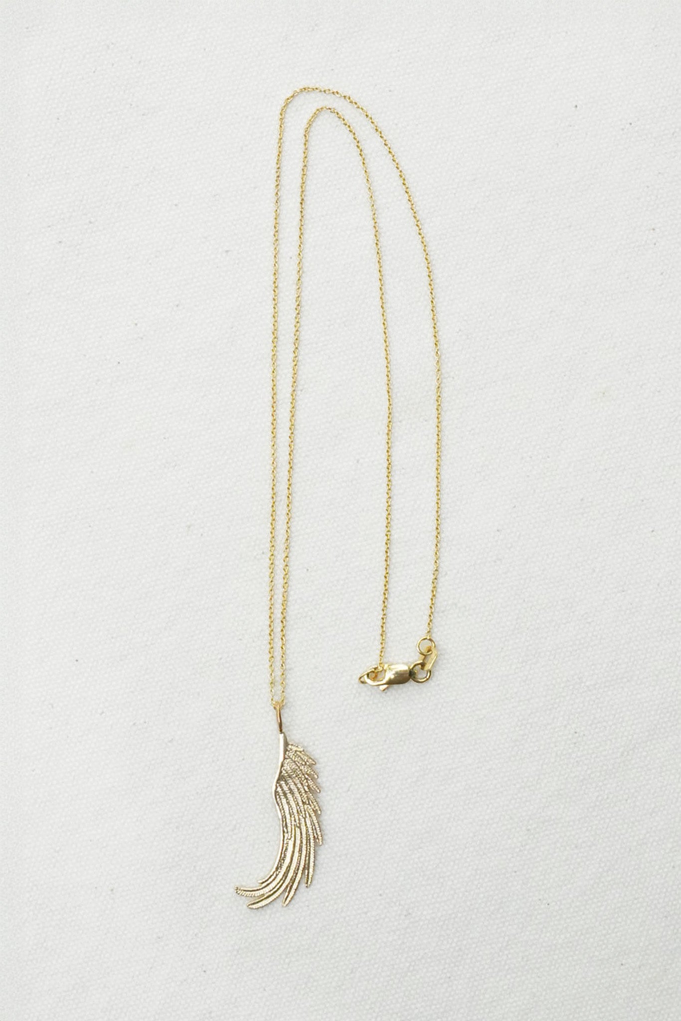 A delicate gold chain necklace featuring a stylized feather pendant and a small bird charm, displayed on a textured off-white background.