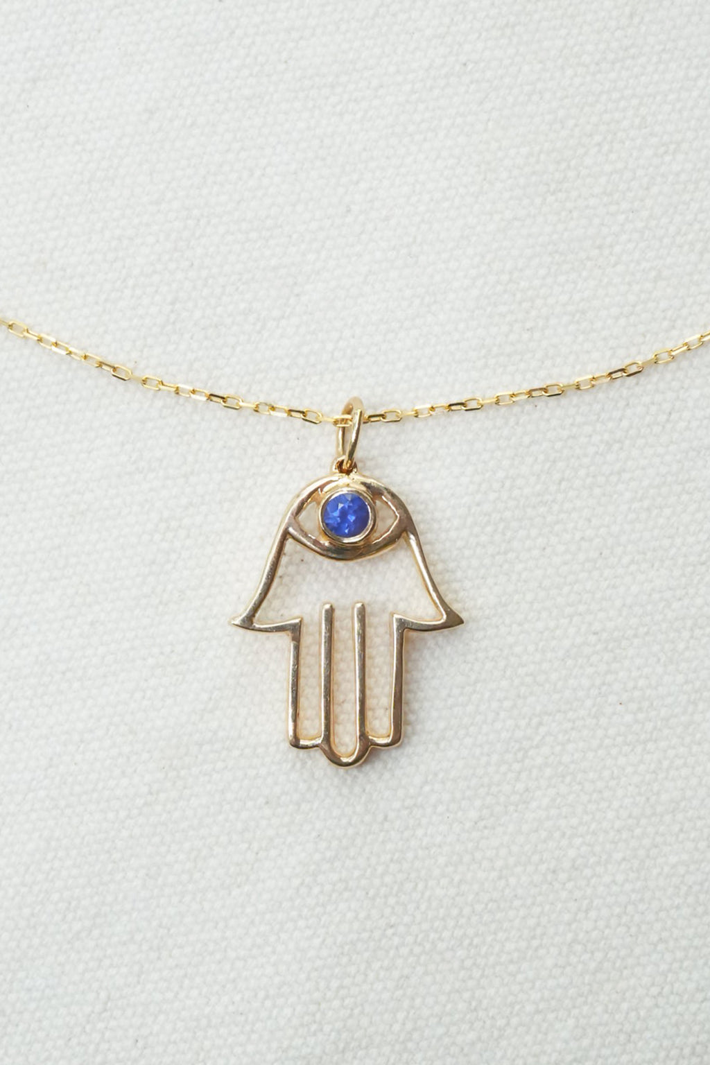 A gold Hamsa hand pendant necklace with a small blue gemstone in the eye, displayed on a textured white background.