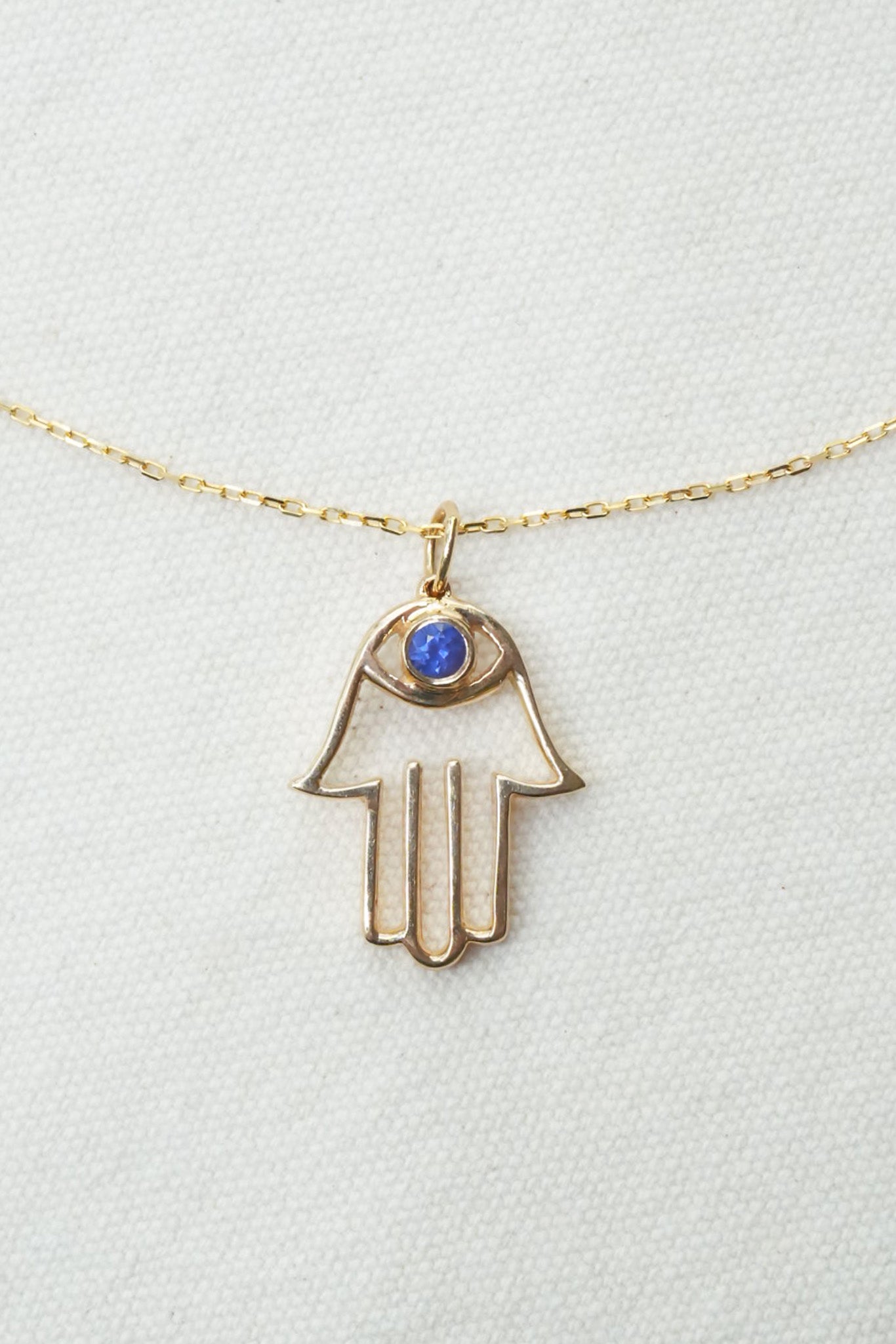 A gold Hamsa hand pendant necklace with a small blue gemstone in the eye, displayed on a textured white background.