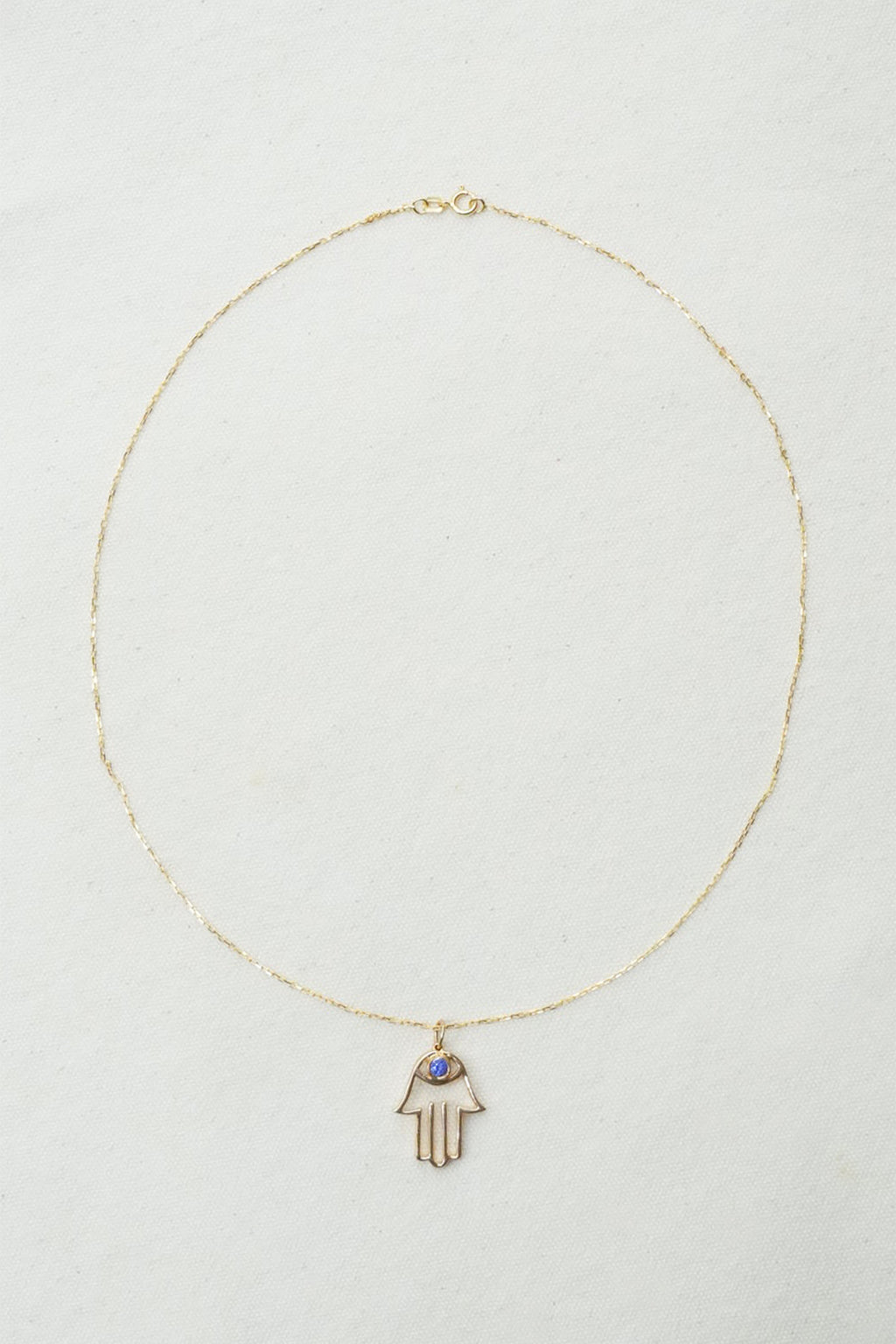 A delicate gold chain necklace featuring a Hamsa hand pendant with a small blue stone accent, displayed on a textured off-white background.