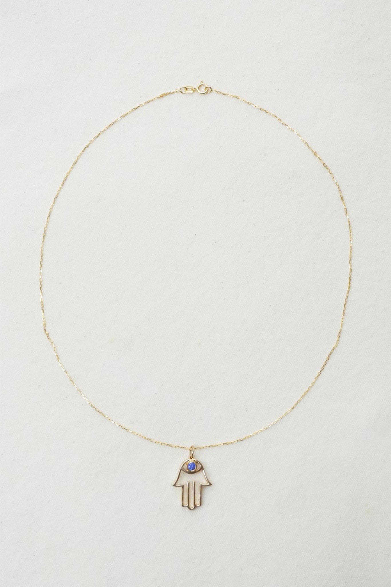 A delicate gold chain necklace featuring a Hamsa hand pendant with a small blue stone accent, displayed on a textured off-white background.