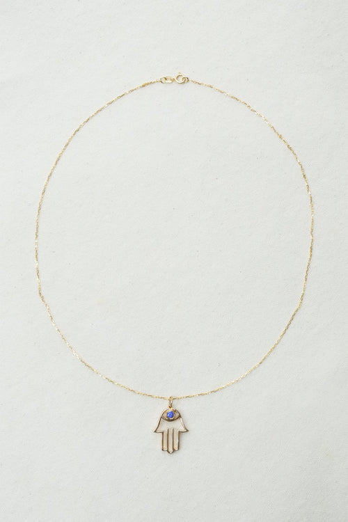 A delicate gold chain necklace featuring a Hamsa hand pendant with a small blue stone accent, displayed on a textured off-white background.