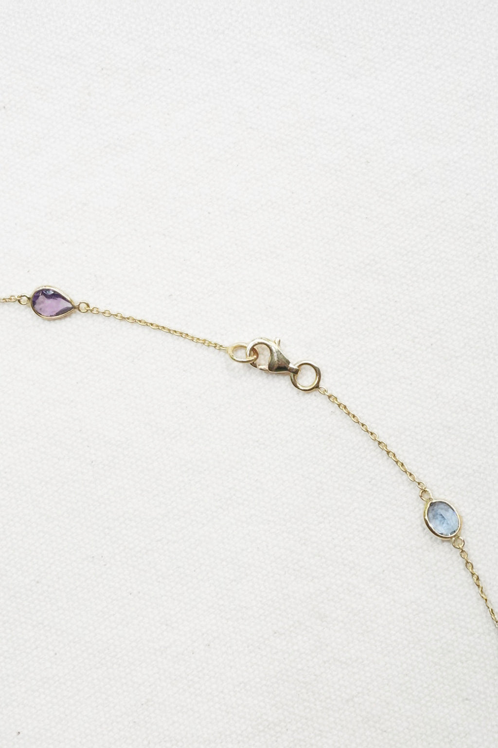 A delicate gold chain bracelet with three small oval gemstone accents in purple, light blue, and clear, displayed on a textured white background.