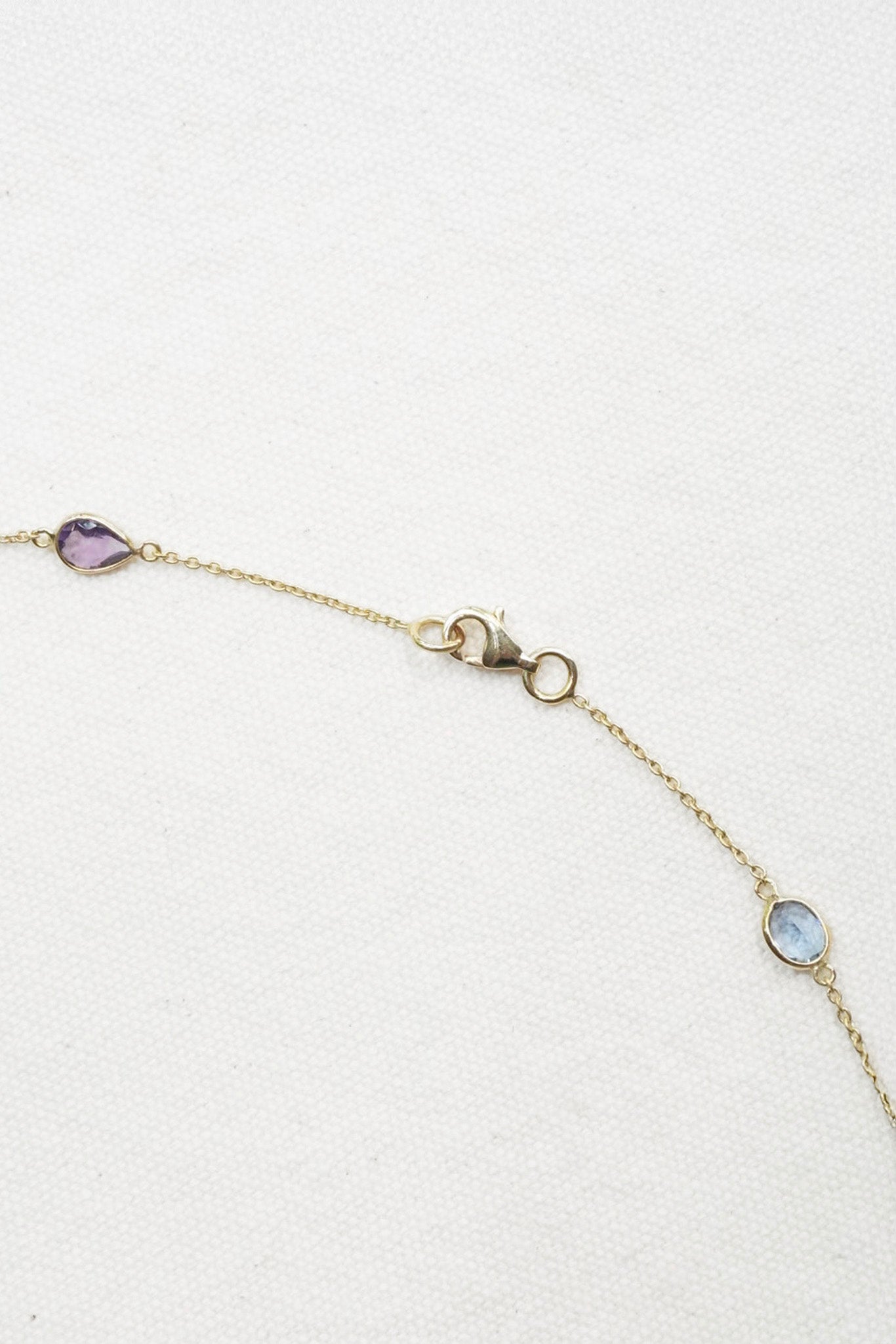 A delicate gold chain bracelet with three small oval gemstone accents in purple, light blue, and clear, displayed on a textured white background.