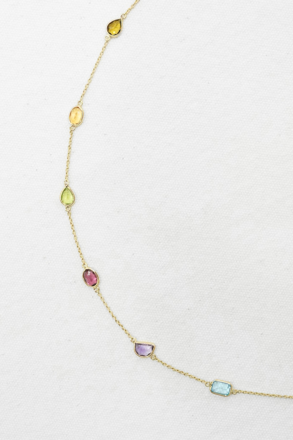 A delicate gold chain necklace featuring seven small, colorful gemstone charms in different shapes and hues, displayed on a textured white background.