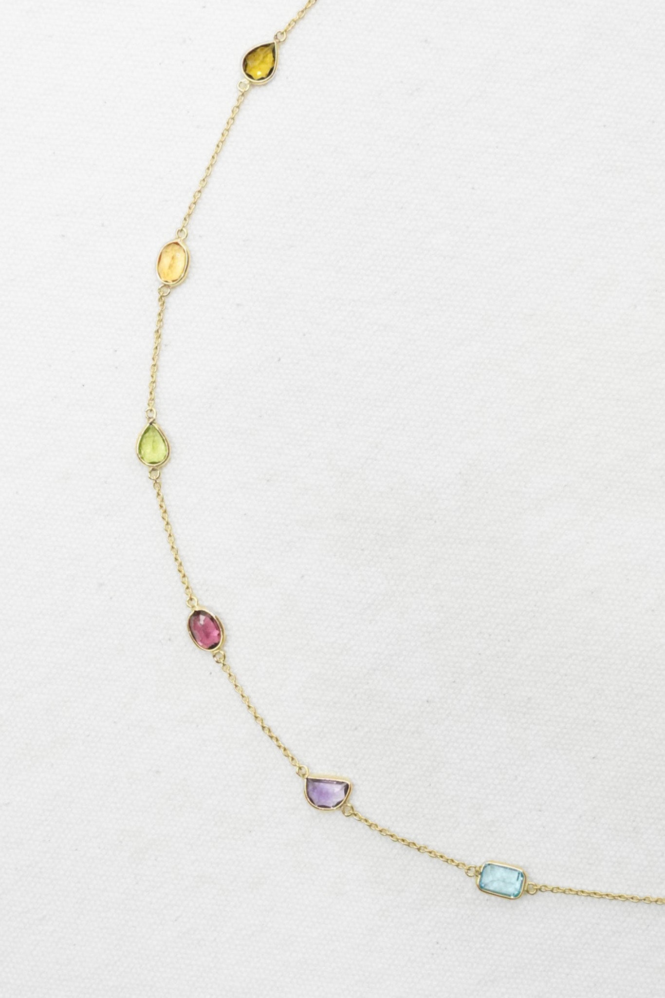 A delicate gold chain necklace featuring seven small, colorful gemstone charms in different shapes and hues, displayed on a textured white background.