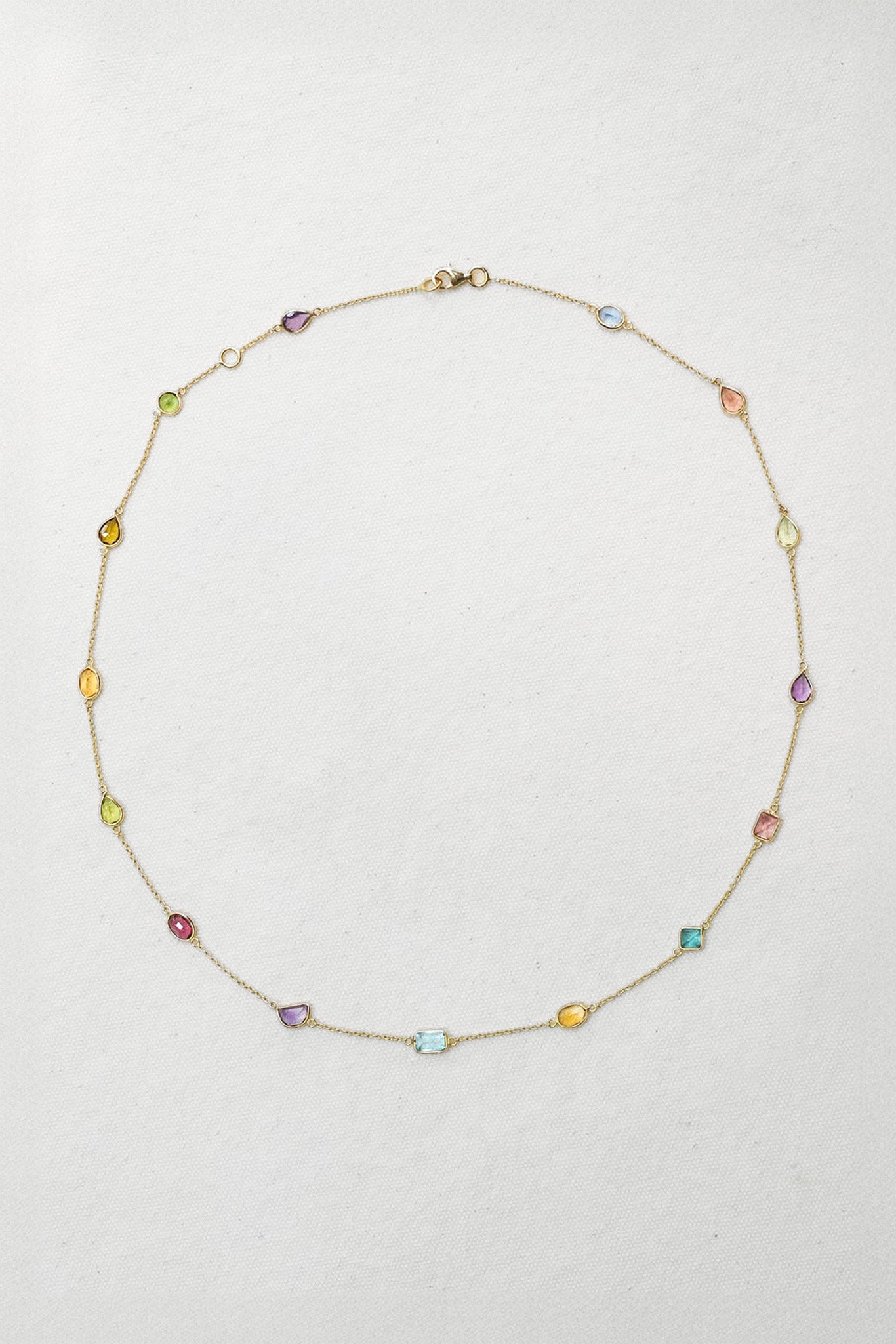 A delicate gold chain necklace with small, colorful faceted gemstone beads spaced along the chain, arranged in a circle on a light textured background.