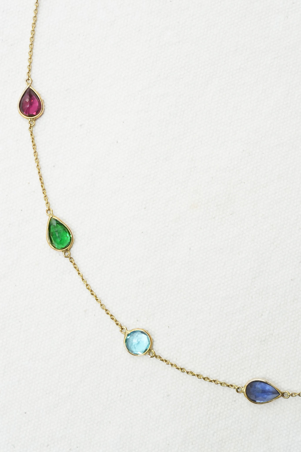 A gold chain necklace featuring five small teardrop-shaped gemstones in pink, green, light blue, and dark blue, arranged diagonally on a textured off-white background.