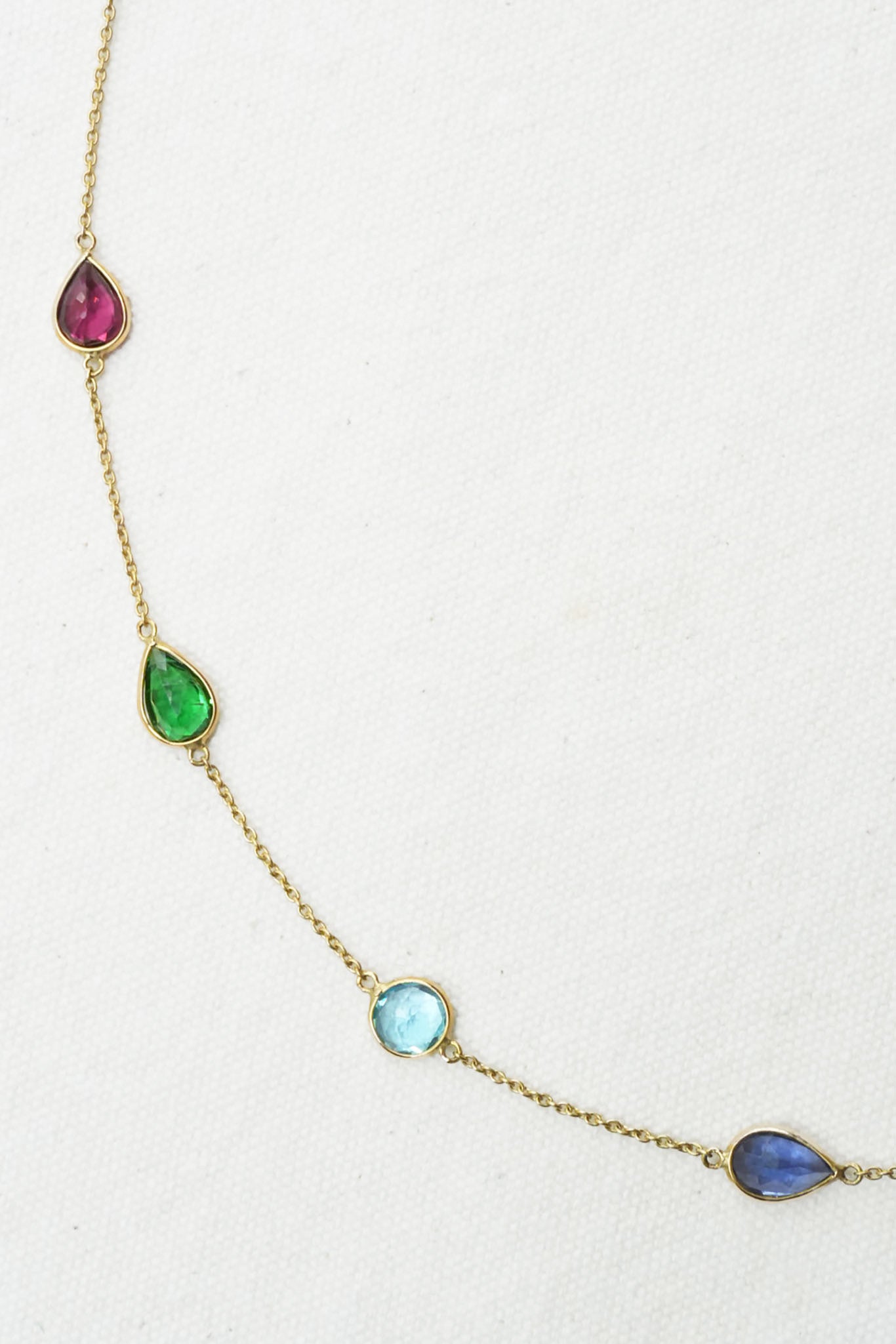 A gold chain necklace featuring five small teardrop-shaped gemstones in pink, green, light blue, and dark blue, arranged diagonally on a textured off-white background.