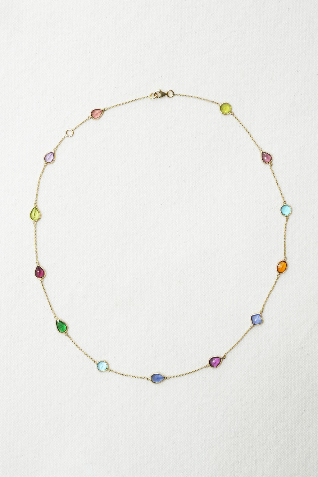 A delicate gold chain necklace featuring multiple small, colorful teardrop-shaped gemstones in various hues, arranged in a circle on a textured off-white background.