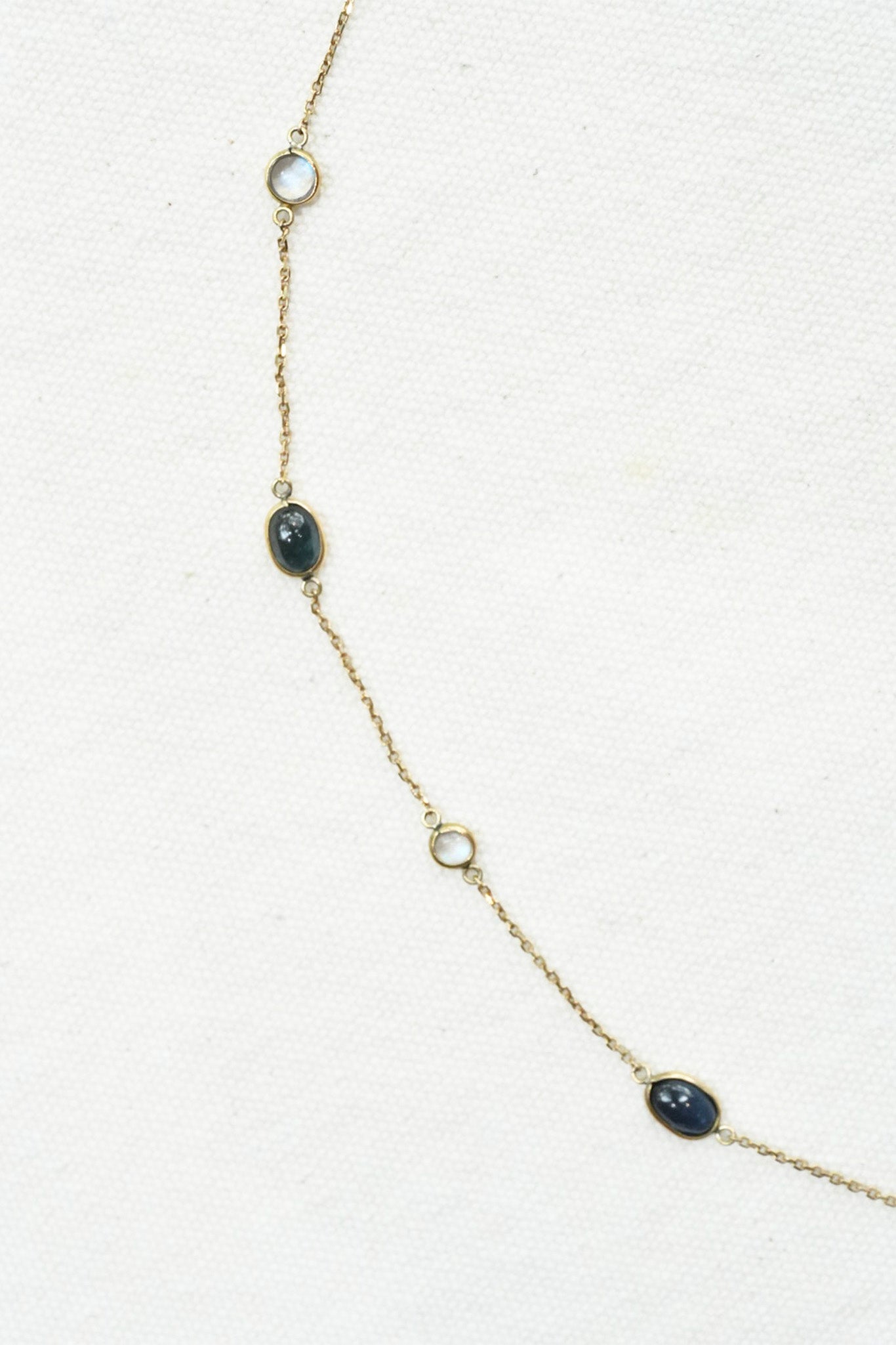 A delicate gold chain necklace featuring three oval dark blue-gray gemstones and two round white stones, displayed on a textured off-white background.