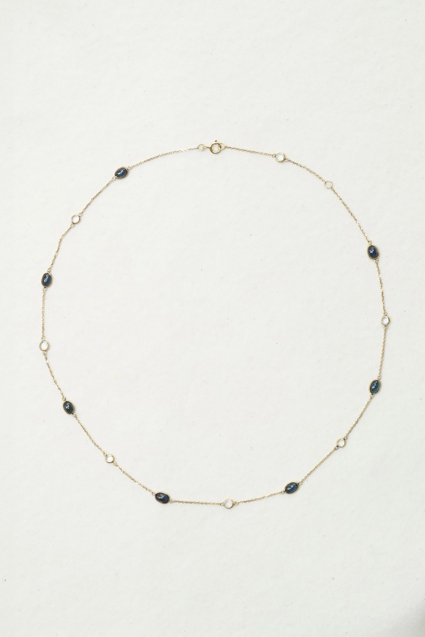 A delicate gold chain necklace with alternating dark oval beads and small clear crystal accents, laid out in a circle on a light off-white background.