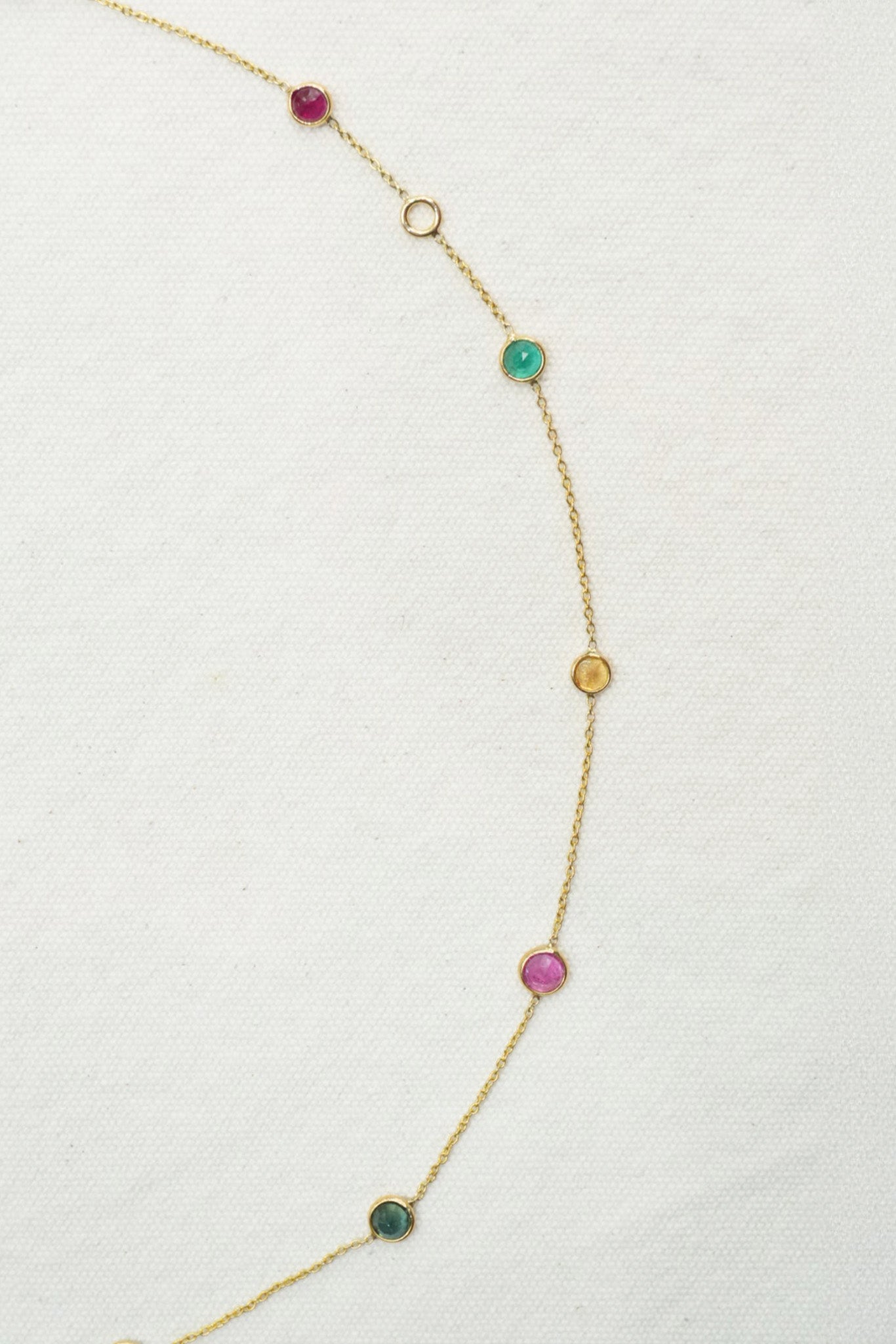 A delicate gold chain necklace featuring multiple small, spaced circular gemstone charms in pink, green, and gold, displayed on a textured off-white fabric background.