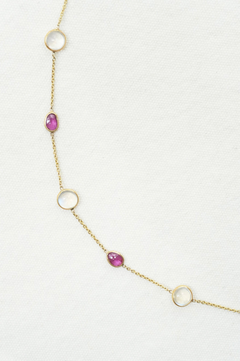 A delicate gold chain necklace featuring alternating open circular loops and small oval bezel-set pink gemstones, displayed on a textured white background.