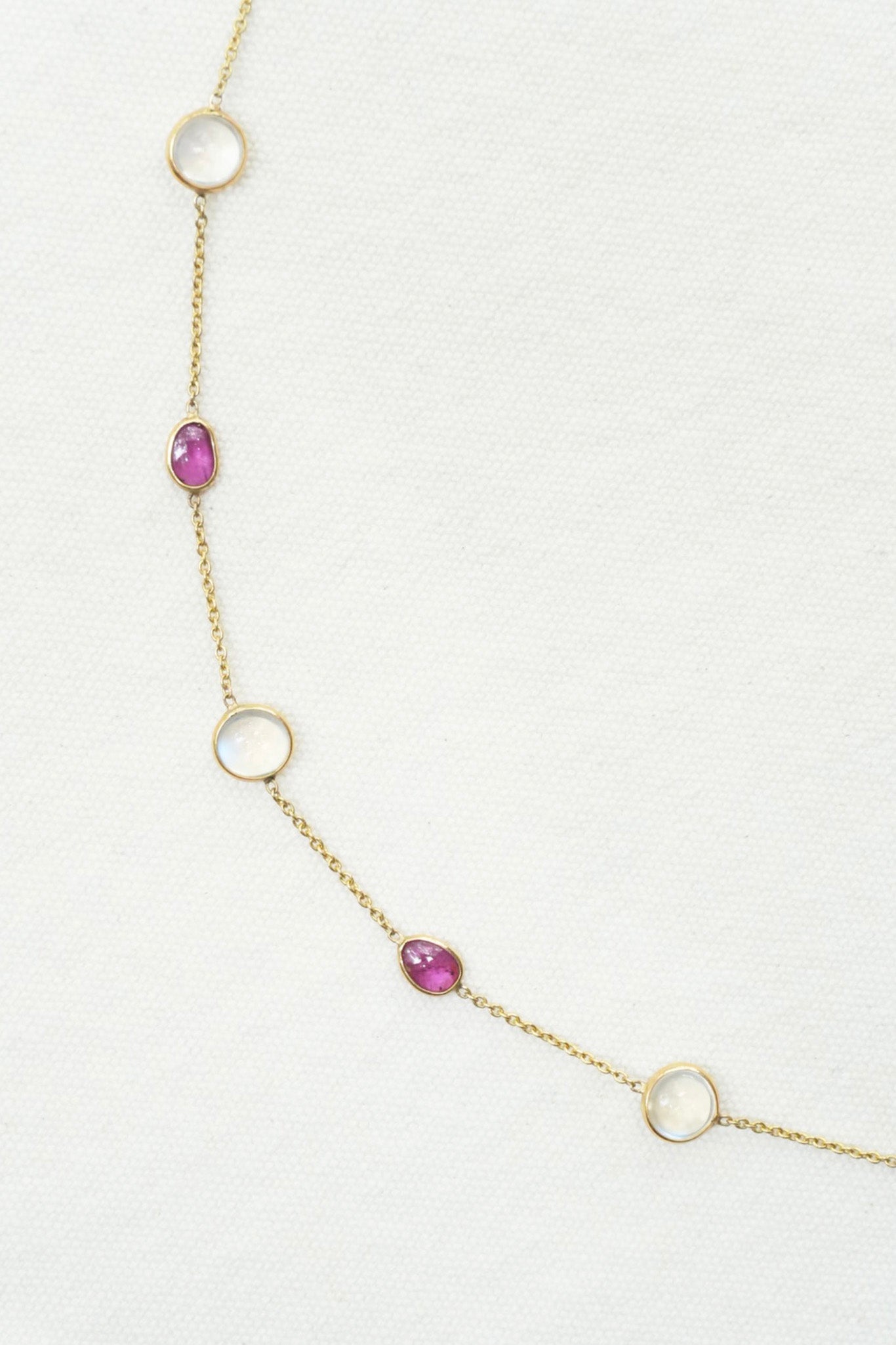 A delicate gold chain necklace featuring alternating open circular loops and small oval bezel-set pink gemstones, displayed on a textured white background.