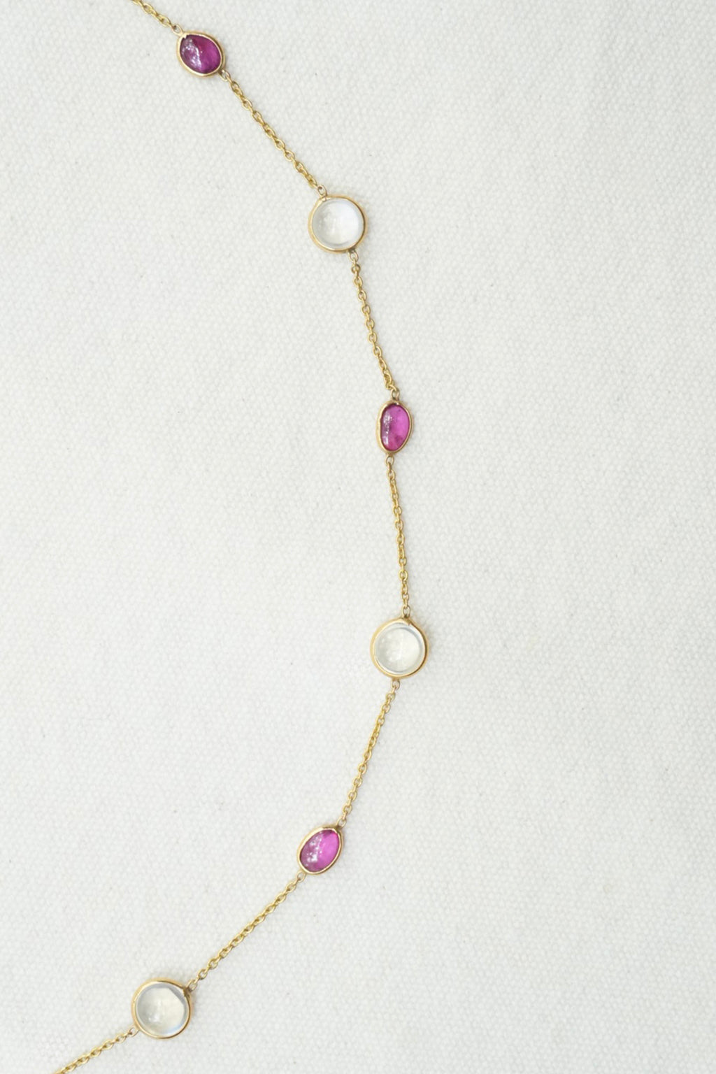 A delicate gold chain necklace featuring alternating round and teardrop-shaped pink gemstones, laid out on a textured off-white background.