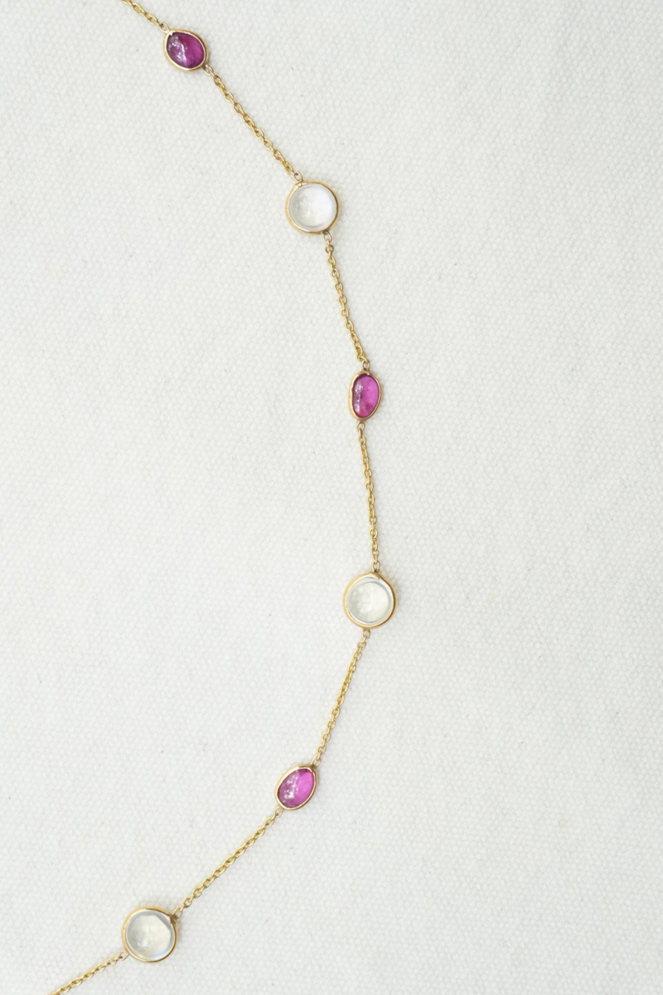 A delicate gold chain necklace featuring alternating round and teardrop-shaped pink gemstones, laid out on a textured off-white background.