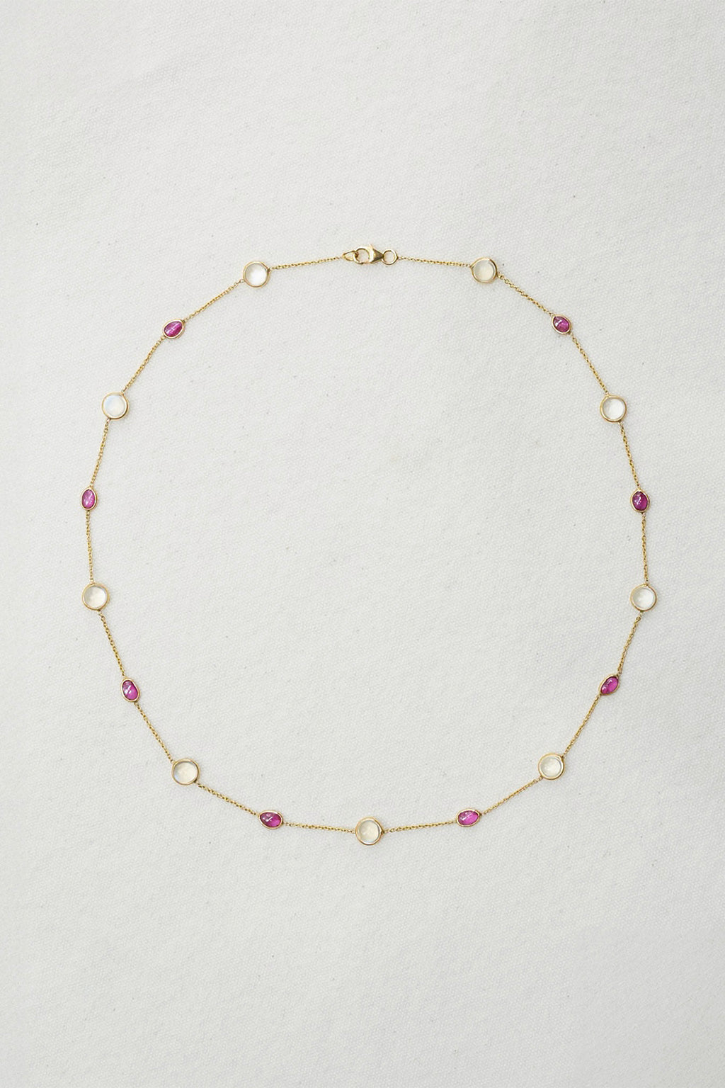 A delicate gold chain necklace featuring alternating round white bezel-set stones and small oval pink gemstones, laid flat on a textured off-white background.