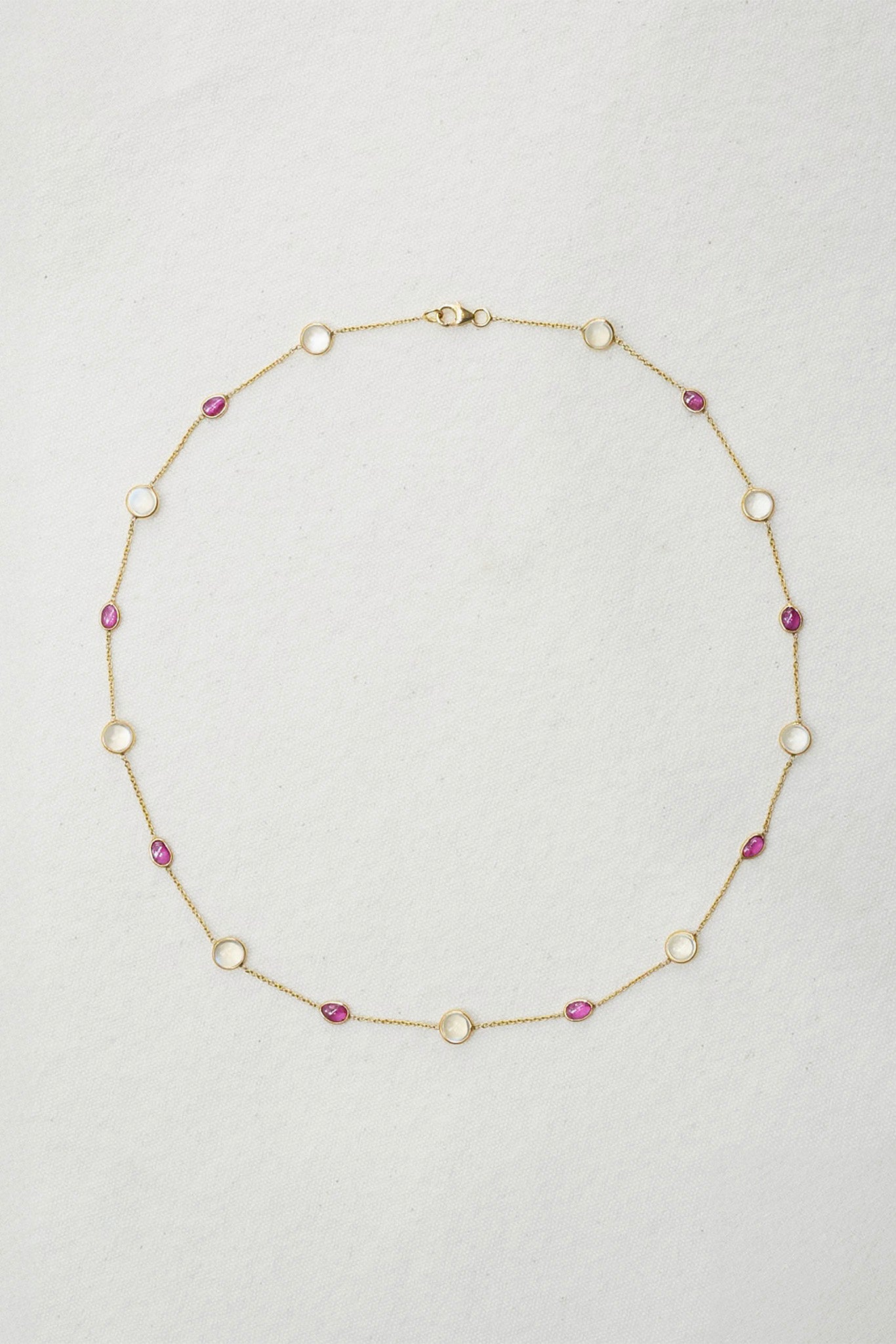 A delicate gold chain necklace featuring alternating round white bezel-set stones and small oval pink gemstones, laid flat on a textured off-white background.