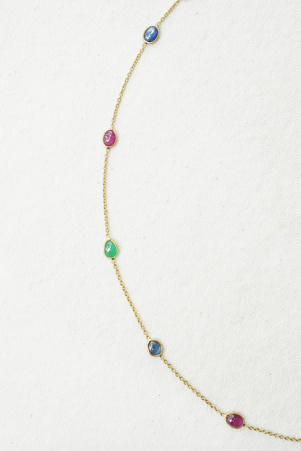 A delicate gold chain necklace featuring five small, evenly spaced oval gemstones in blue, pink, green, blue, and pink, laid out on a textured off-white background.