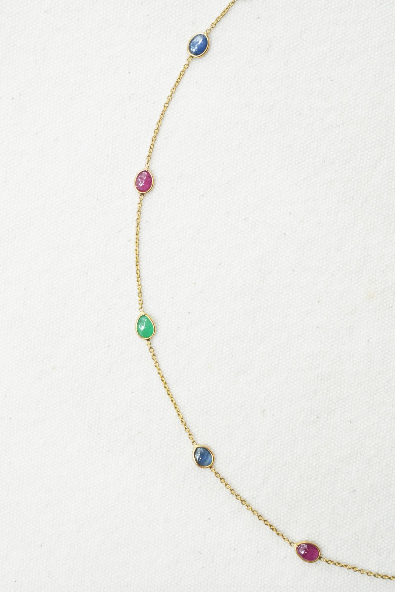 A delicate gold chain necklace featuring five small, evenly spaced oval gemstones in blue, pink, green, blue, and pink, laid out on a textured off-white background.